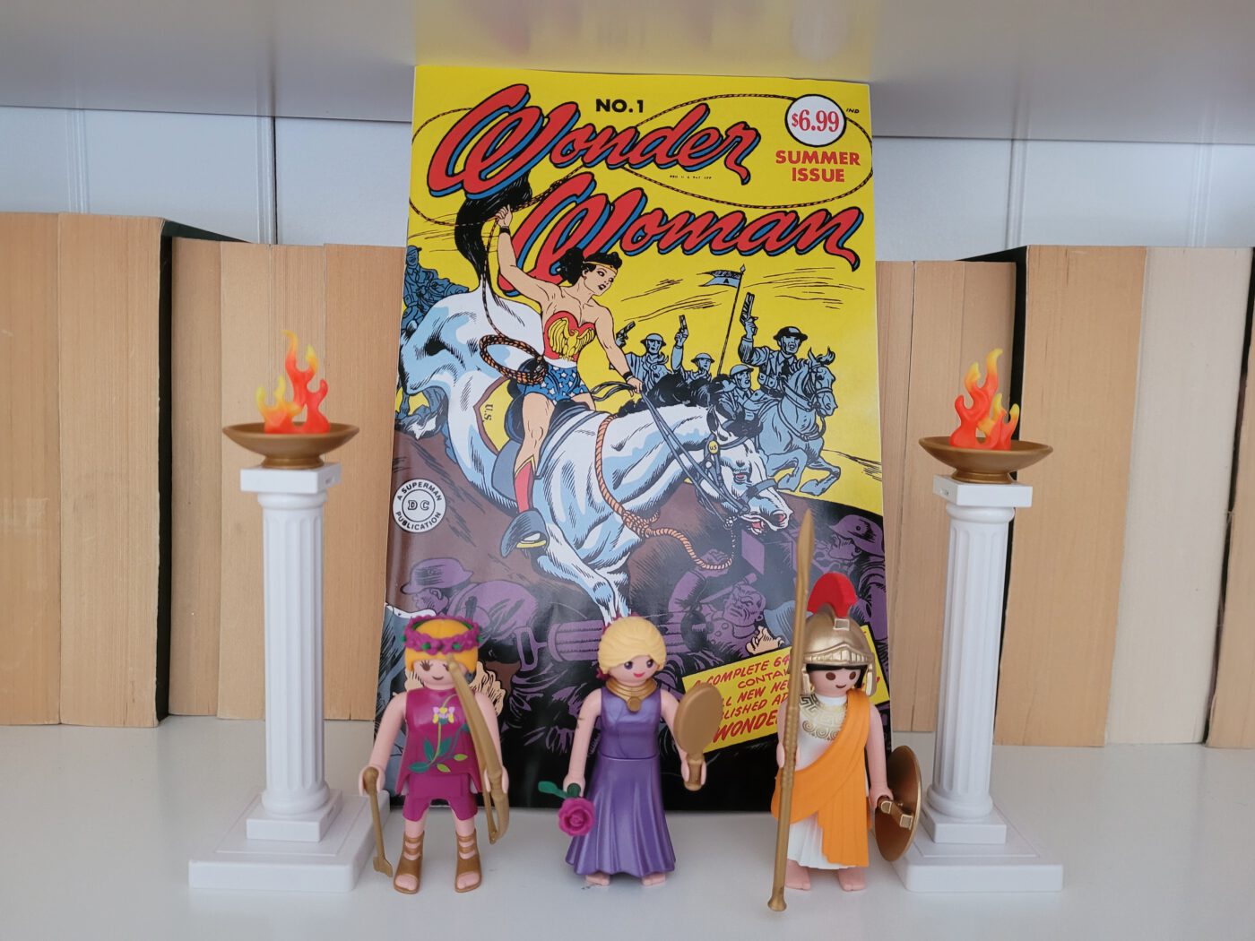 Das Bild zeigt einen Nachdruck des ersten Wonder Woman-Hefts in meinem Bücherregal. Davor stehen Playmobilfiguren der Göttinnen Artemis, Aphrodote und Athene